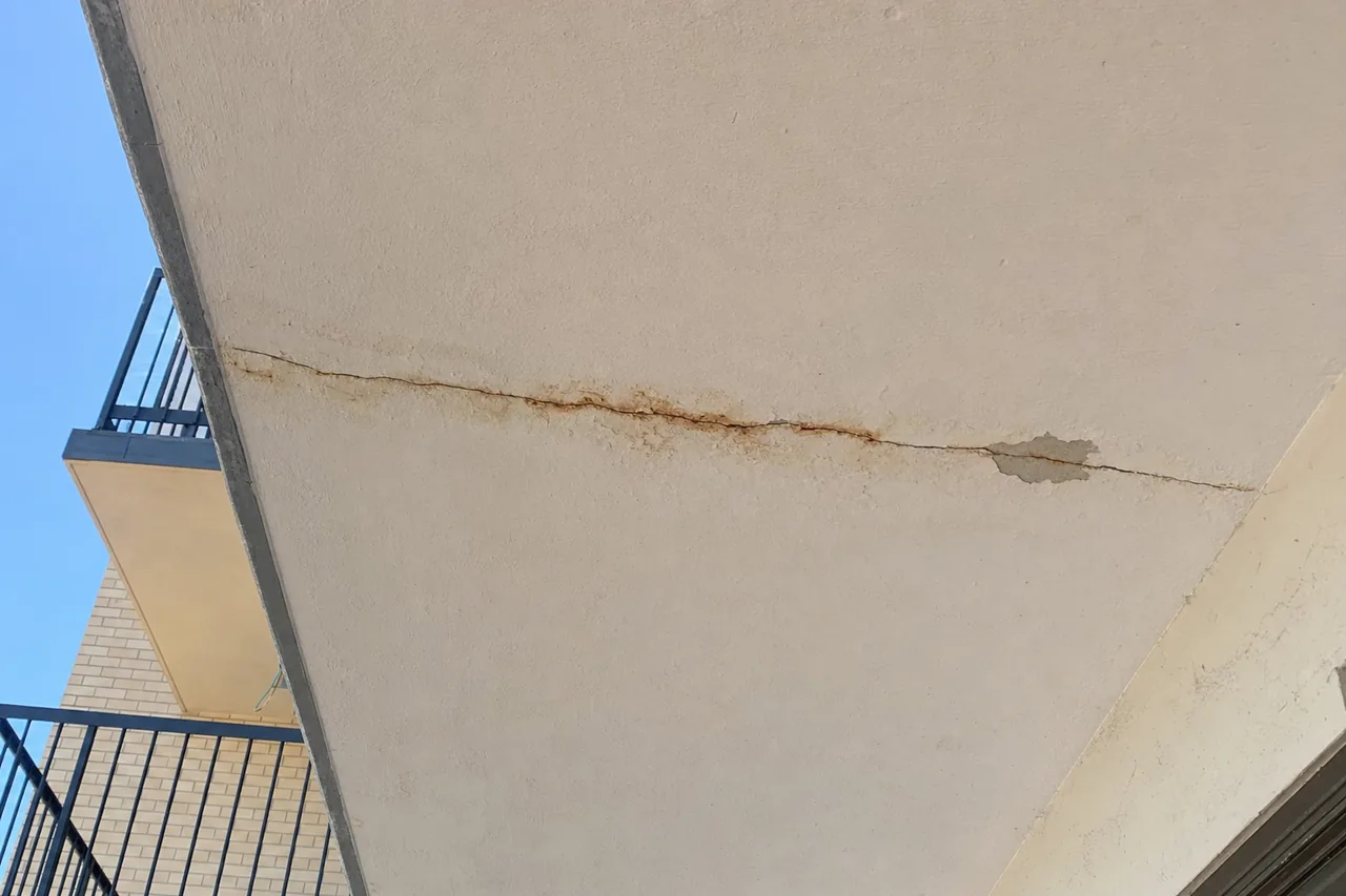 The underside of a concrete apartment balcony with rust-brown staining bleeding through flaking cream paint along a hairline crack, and a small patch of paint lifting beside it — concrete cancer repair, Brisbane. Illustrative example.